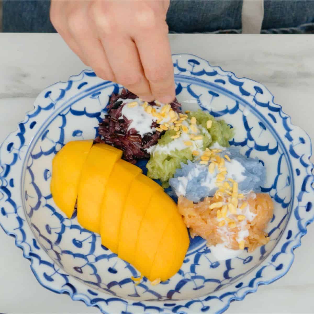 a plate of mangoes with 4 colours of sticky rice, and coconut sauce on top, with mung beans being sprinkled on the rice.
