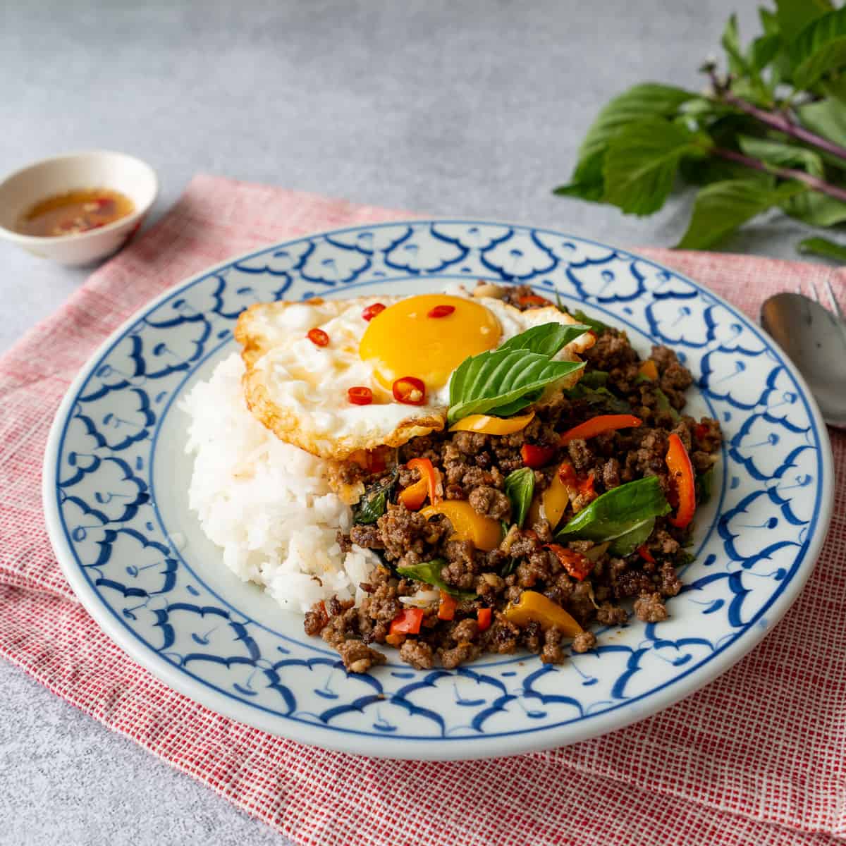 a plate of pad gaprao beef with rice and a fried egg on top, with a bowl of fish sauce on the side.