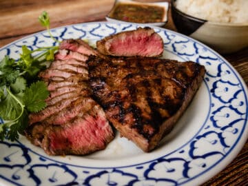 A plate of grilled steaks, some thinly sliced with dipping sauce and sticky rice on the side.