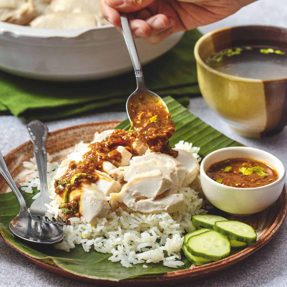 a plate of Hainanese chicken rice with sauce being drizzled on it. A bowl of broth on the side.