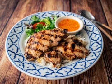 A plate of grilled lemongrass chicken on rice with a side of nuac cham dipping sauce and salad greens.