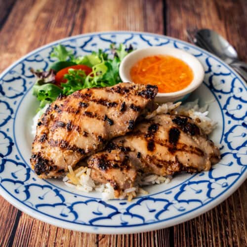 A plate of grilled lemongrass chicken on rice with a side of nuac cham dipping sauce and salad greens.