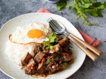 a plate of pad kra pao beef with rice and a fried egg. A side of fish sauce condiment and holy basil sprigs
