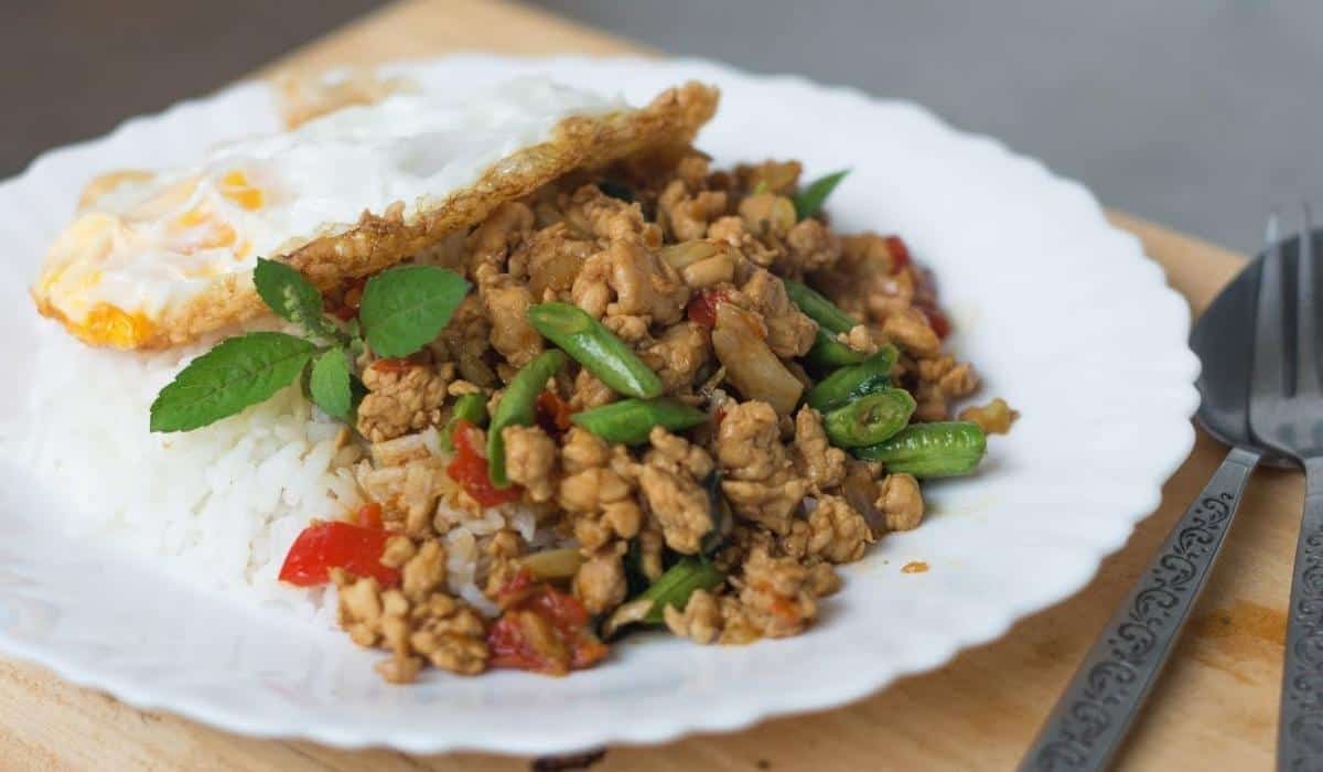 A heaping serving of pad gaprao lay on a bed of rice and topped with a beautiful over-easy fried egg. Garnished with a sprig of herb, the dish is decorated with splashes of green and red from the pad gaprao's veggie mix. The plate is placed on a wooden mat with a metal spoon and fork set to the side.