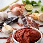 A bowl of red curry paste with ingredients in the background. Text overlay: Thai red curry paste Easy vs Traditional Way.