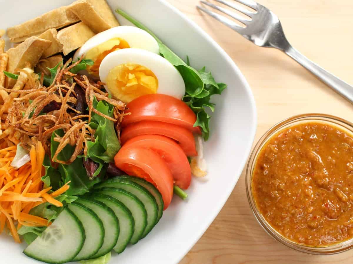 A bowl of salad veggies with a boiled egg with a bowl of peanut dressing on the side.