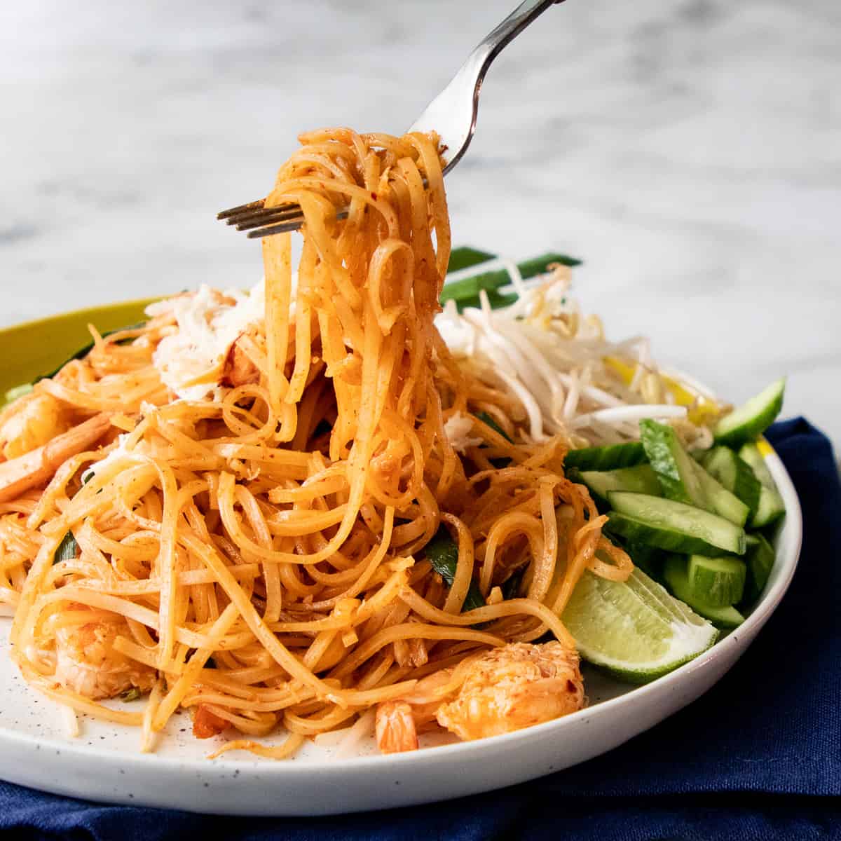 a plate of sen chan pad pu with a fork lifting up noodles with Cucumber, beansprouts and a lime wedge on the side
