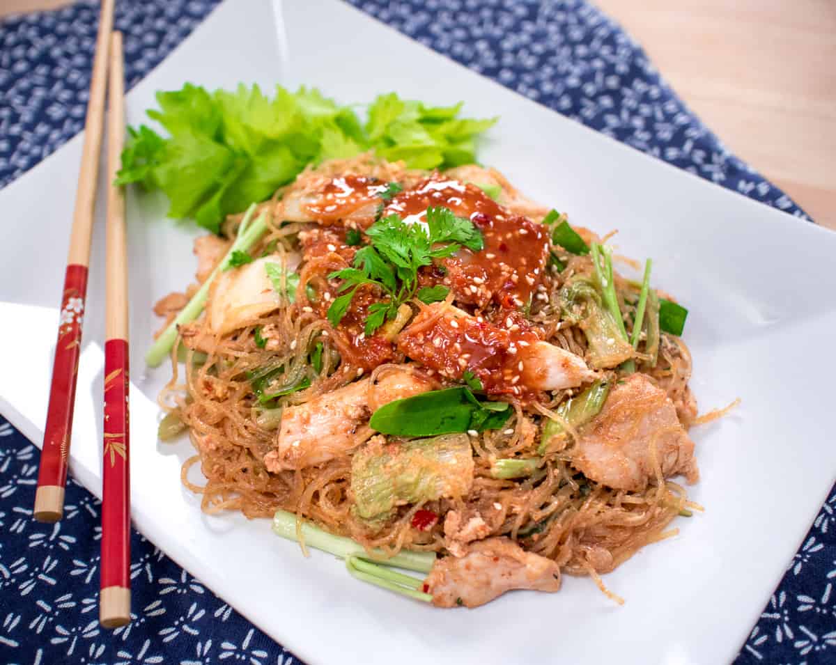 A generous portion of Suki Hang is placed on a white square plate. The celery brings a bright green composition to the over all look as the red sauce glistens on top of the delicious slices of chicken. 