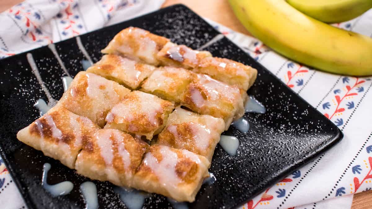 Placed on a black square plate, a crispy Banana roti is perfectly cut into 12 pieces. Sweetened condensed milk is drizzled on top with a light powdering of icing sugar.