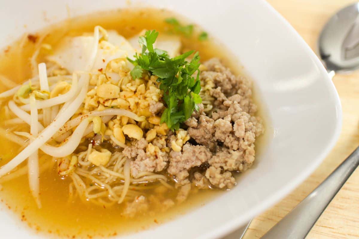 A golden steaming bowl of tom yum noodles in a white bowl. Topped with vilantro and beansprouts.