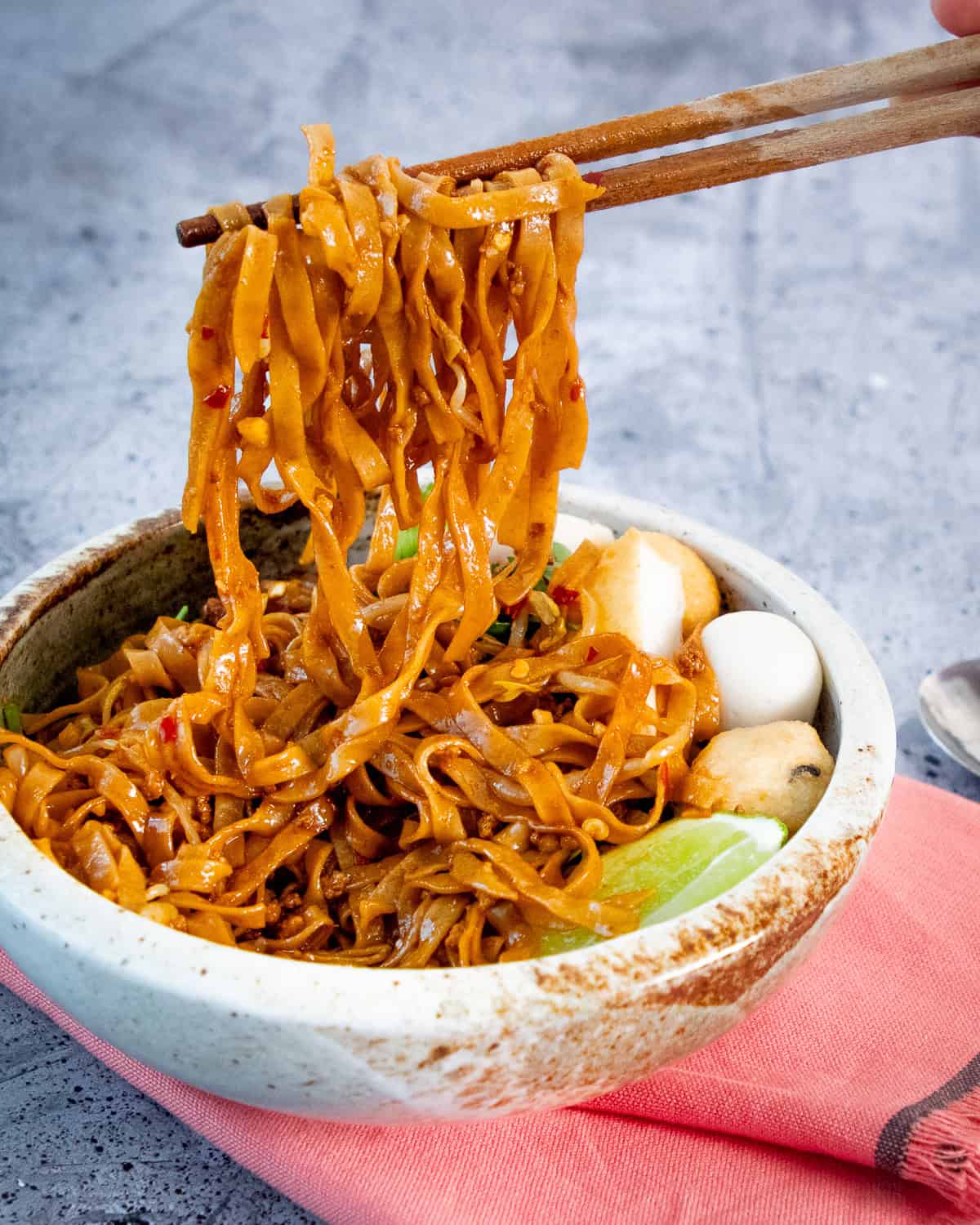 Chopsticks holding up tossed noodles in a bowl.